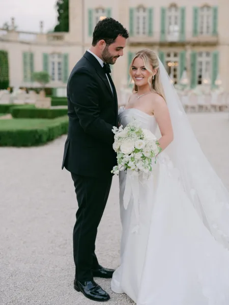 Couple portrait, an outdoor wedding portrait with the groom gazing at his bride as she holds a white bouquet, before an estate with green shutters.