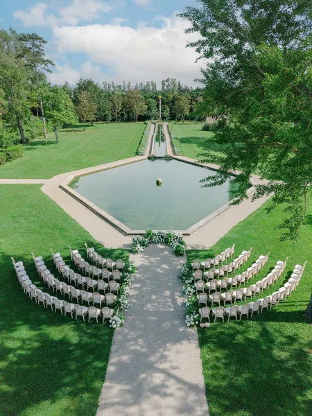 Outdoor ceremony setup with garden ceremony decor, white florals edging curved ceremony seating beside a reflecting pool on a tree-lined lawn.