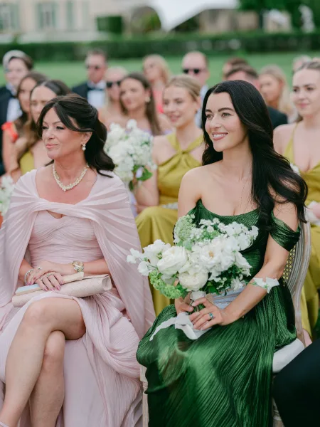 Ceremony guests showcase wedding guest style, one draped in a shawl, seated on a garden lawn framed by hedges and a grand building facade.