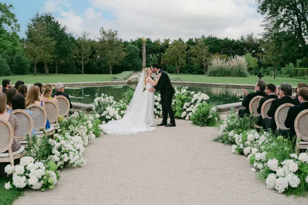 Wedding first kiss at an outdoor wedding ceremony, the bride's long veil trailing as the couple embraces by a reflecting pool amid garden trees.