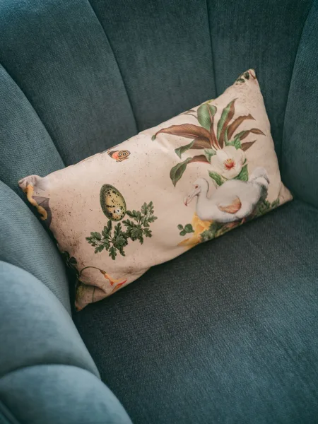 Throw pillow styled as a botanical lumbar pillow, showcasing a swan motif, set on a teal upholstered chair with channel tufting.