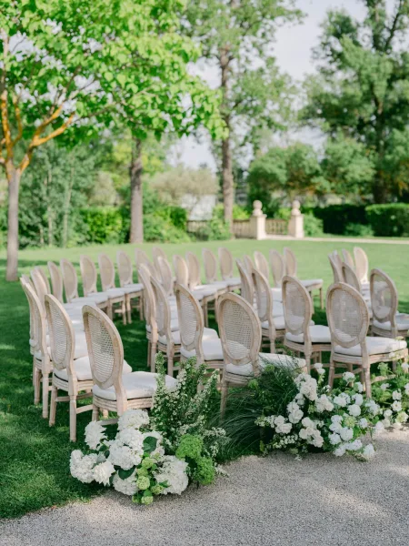 Ceremony aisle design for a garden ceremony seating area featuring cane-back chairs along a gravel path framed by hedges and a stone balustrade.