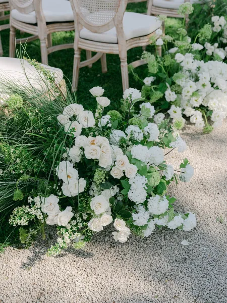 Ceremony aisle flowers define garden ceremony decor, with white roses clustering low along a gravel path on a grass lawn.