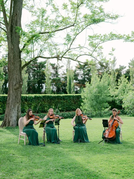 String quartet wedding ceremony musicians in green dresses perform on a grass lawn beneath a large tree, with hedges and open sky beyond.