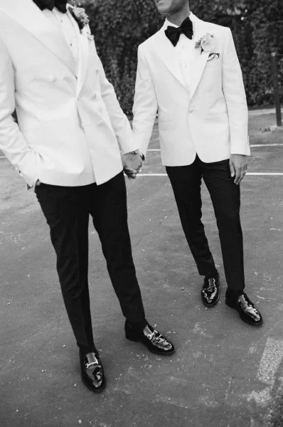 Groom portrait in a white dinner jacket with black bow tie and boutonniere, standing on pavement beside a hedge wall outdoors