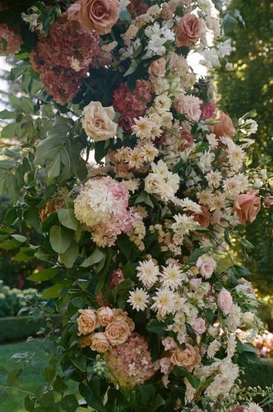 Wedding floral installation with rose blooms and hydrangea, white dahlias and greenery on a ceremony pillar in a sunlit garden backdrop