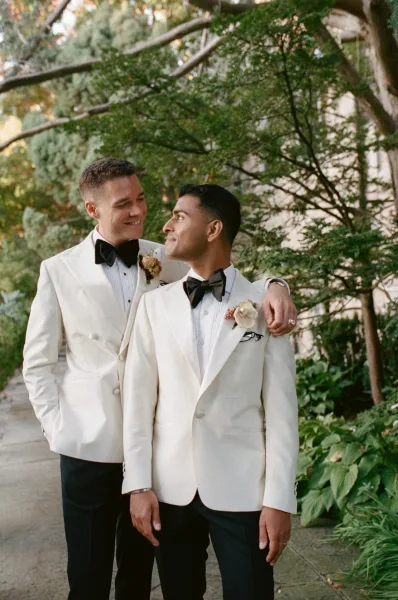 Groom portrait in a white tuxedo jacket with black bow tie and boutonniere, standing on a stone walkway amid garden greenery and trees