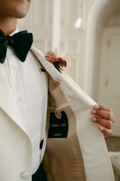 Groom portrait in a white dinner jacket with a black bow tie, holding his lapel to show boutonniere against a soft neutral backdrop