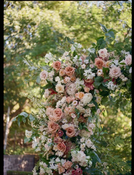 Wedding ceremony florals on a floral ceremony arch with blush and peach roses, white hydrangea, and greenery in sunlit garden bokeh
