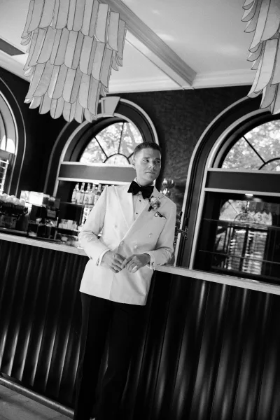 Groom portrait in a white tuxedo groom look, leaning on a bar with black bow tie, boutonniere, and liquor bottles behind him
