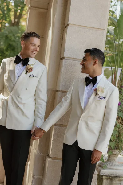 Groom portrait of two same sex grooms holding hands in white dinner jackets with boutonnieres, standing by stone columns in a garden courtyard