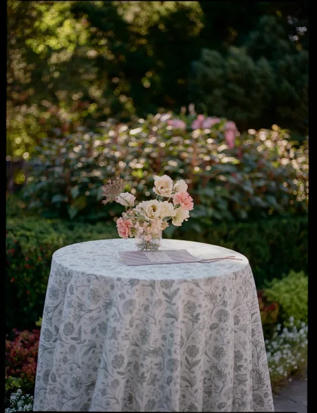 Welcome table decor with a wedding guestbook table, ribbed glass vase of blush-and-white flowers on patterned linen, set amid garden greenery