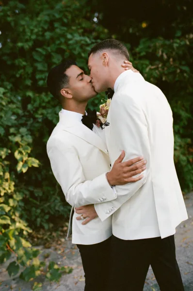 Wedding kiss portrait of two grooms in matching white tuxedos and black bow ties, embracing on a garden pathway with lush greenery backdrop
