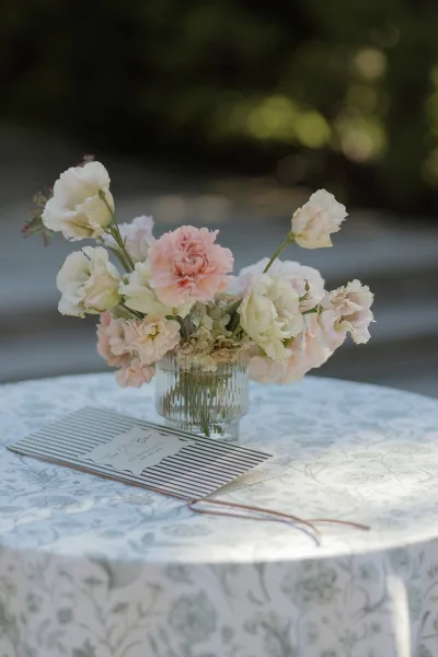 Invitation suite with striped wedding envelope and ribbon beside a glass vase of pink and white flowers on a patterned tablecloth outdoors
