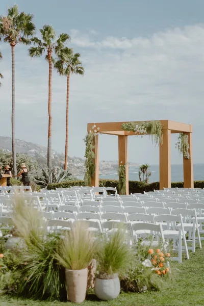 Ceremony setup with outdoor ceremony seating facing a wooden arch, framed by palm trees with the ocean and distant hills beyond.