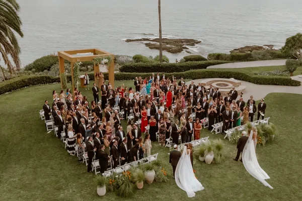 Ceremony processional with brides walking down the aisle beneath a wooden chuppah, ocean and palm trees beyond.