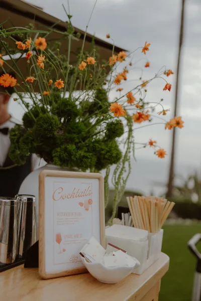 Signature cocktail sign for a wedding cocktail menu, trimmed with orange flowers, with an ocean view and palm trees in the background.