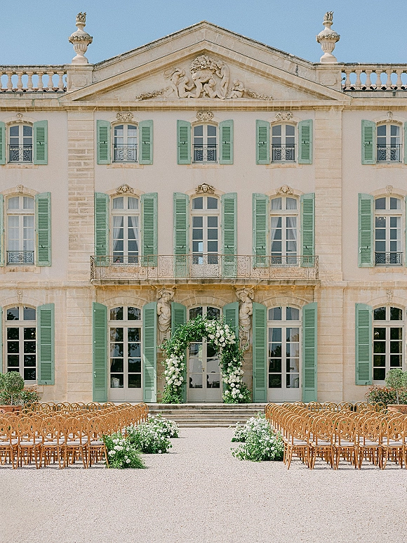 Ceremony setup with a floral arch of white flowers and greenery, wooden chairs lining a gravel aisle before a stone villa courtyard