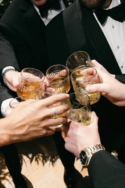 Groomsmen toast as the wedding party cheers, hands meeting with whiskey glasses, cast shadows stretching across a tile floor.
