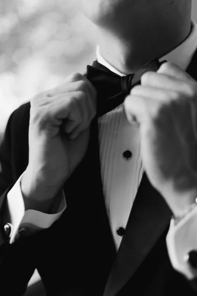 Groom getting ready, tying a bow tie with steady hands as he adjusts his crisp shirt, a focused moment before the ceremony.