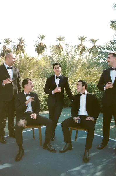 Groomsmen portrait of a groom with groomsmen, raising a whiskey glass, posed under palm trees and greenery beneath a clear sky on a paved path.