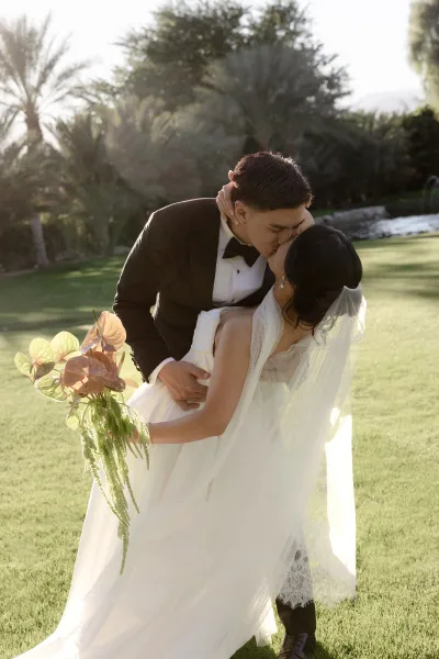 Wedding kiss portrait, with groom dipping bride beside a palm-lined pond and fountain, her veil flowing.