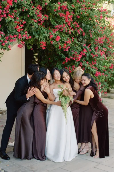 Bridal party portrait of a bride with bridesmaids sharing a warm hug, her cascading bouquet framed by bougainvillea along a stucco wall.