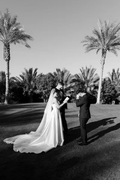 Wedding vows shared as the bride reads from a vow book to her groom beneath palm trees on a grass lawn under a clear sky.
