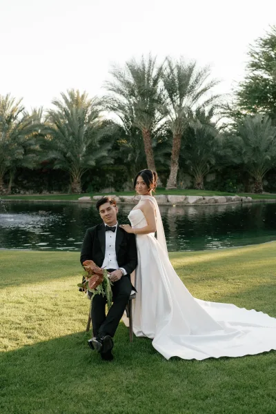Couple portrait of a bride in a long train wedding dress beside her groom by a palm-lined pond and grass lawn under open sky.