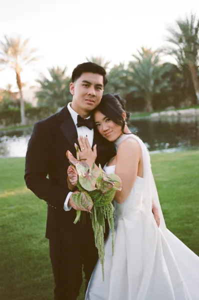 Couple portrait of a bride and groom embracing, the bride holding a bouquet by a pond framed with palm trees.