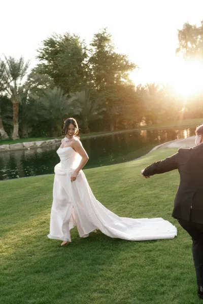 Bridal portrait of a bride laughing with a flowing dress train on a grass lawn beside a pond framed by trees and wide sky.