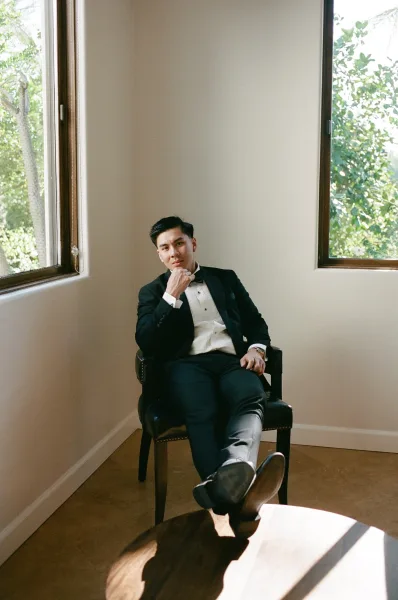 Groom portrait in black tie groom style, seated in an armchair by sunlit windows with greenery beyond and white walls brightening the room.