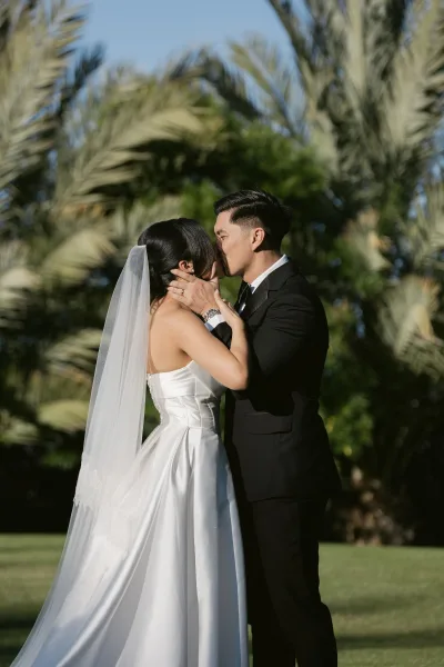 Wedding kiss portrait of a bride in strapless gown and her groom under swaying palm trees and blue sky, on a sunlit garden lawn