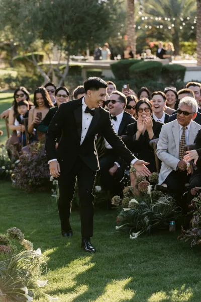 Ceremony processional with groom walking down aisle in a black tuxedo, guests lining a grass lawn beneath palm trees and hedges.