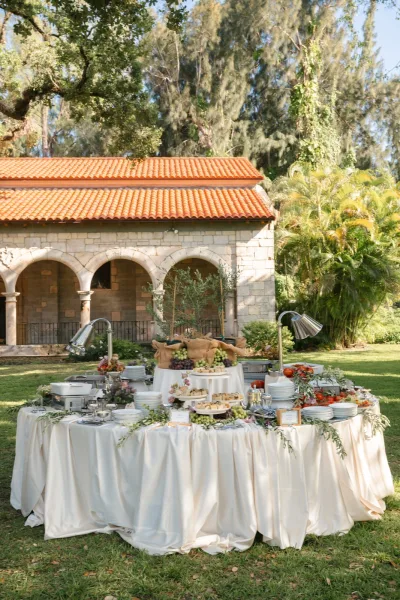 Wedding buffet table serving as a reception food station, styled with olive branches on a grass lawn beside stone arches and palm trees.