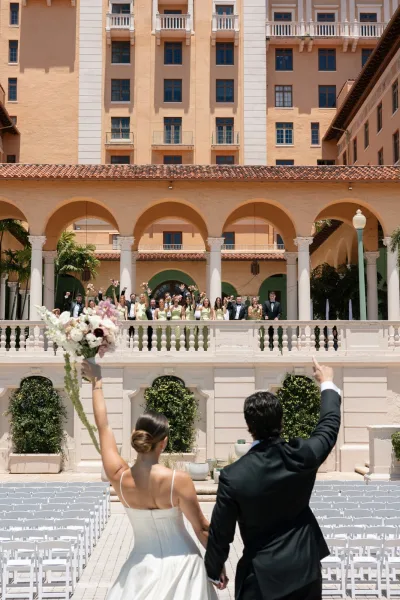 Ceremony exit with the couple waving to guests, the bride lifting her bouquet, in a sunlit courtyard of arches, columns, and palms.