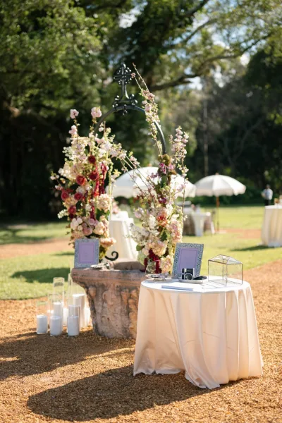 Guest book table styled as an instant camera guest book with a glass card box, set in a garden with trees, lawn, and white umbrellas.