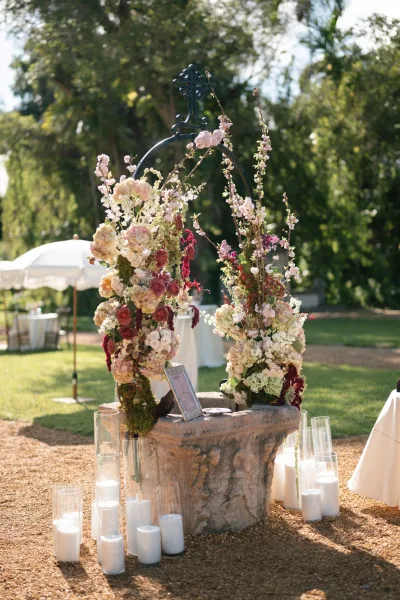 Ceremony flowers frame an iron arch as lush outdoor wedding flowers shape a romantic aisle beneath garden trees and a white umbrella.