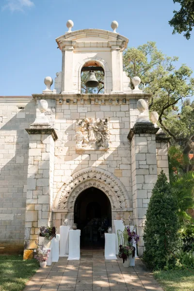 Ceremony entrance decor with arrangements on pedestals at a chapel wedding entrance, before a stone chapel facade and bell tower.