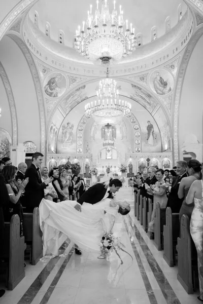 Wedding recessional dip-kiss as newlyweds pause in the church aisle, guests applauding beneath frescoed vaults and framed by altar florals.