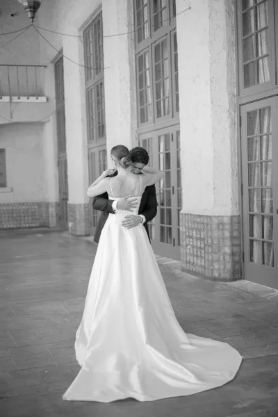 Bride and groom hug in a wedding first look, her gown train trailing as they embrace by French doors in a stucco corridor.