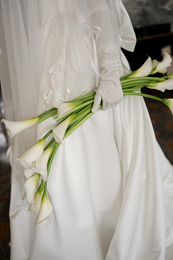 Calla lily bouquet held in white bridal gloves, long green stems tied with satin ribbon bows against a dark indoor background