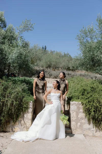 Bride and bridesmaids in olive bridesmaid dresses stand on stone steps amid olive trees, the bride holding a small green bouquet under a blue sky.