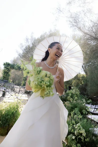 Bridal portrait of a bride with parasol, laughing along a sunlit garden aisle as white ceremony chairs frame the path beneath leafy trees.