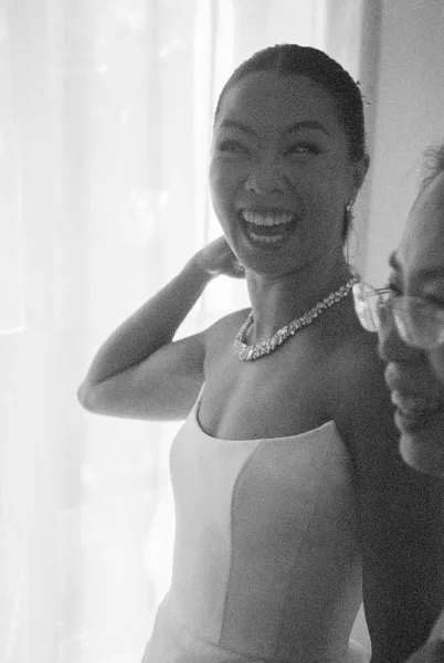 Bridal portrait of a bride getting ready, softly lit by sheer curtains, wearing a delicate necklace and smiling indoors.