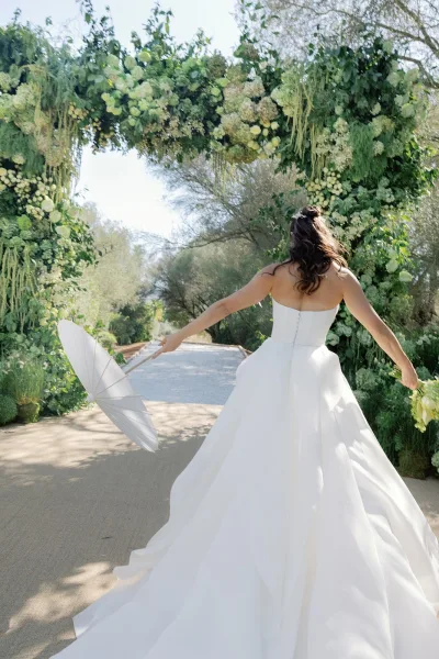 Bridal portrait of a bride with parasol, walking along a sunlit garden path framed by trees and greenery, captured in a candid, backlit moment.