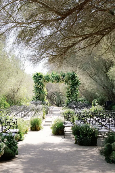 Ceremony setup with a floral wedding arch along a concrete path in a garden beneath a woodland canopy.