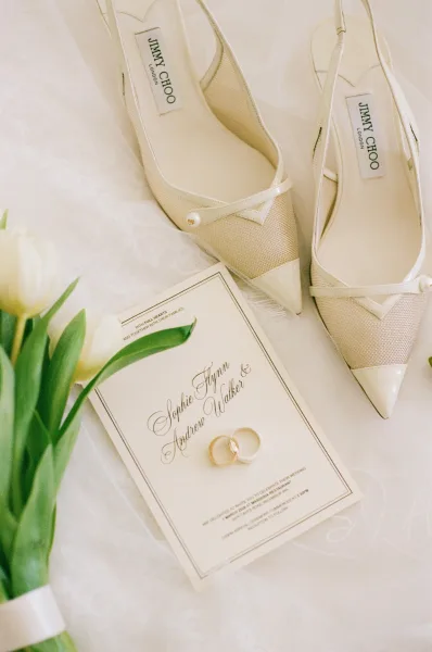 Bridal shoes, pointed toe wedding heels with pearl embellishment, rest on lace fabric and a soft tulle veil in a light, airy flatlay.