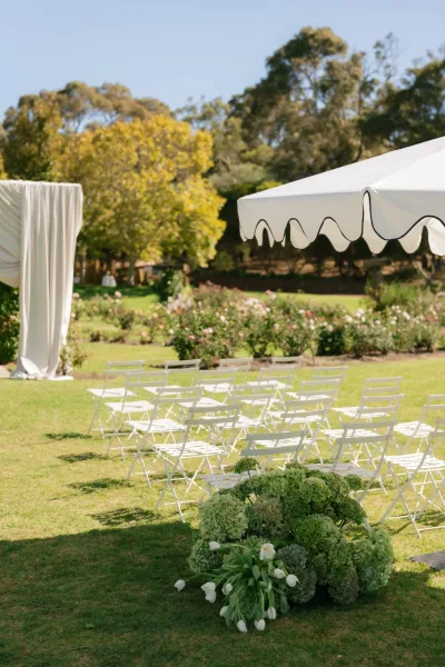 Ceremony setup with outdoor ceremony seating in rows of white folding chairs on a grass lawn framed by garden trees and rose bushes.
