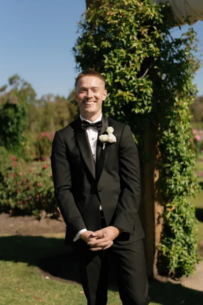 Groom portrait of a black tie groom with a single boutonniere, standing by a vine-covered garden arbor under a clear blue sky.